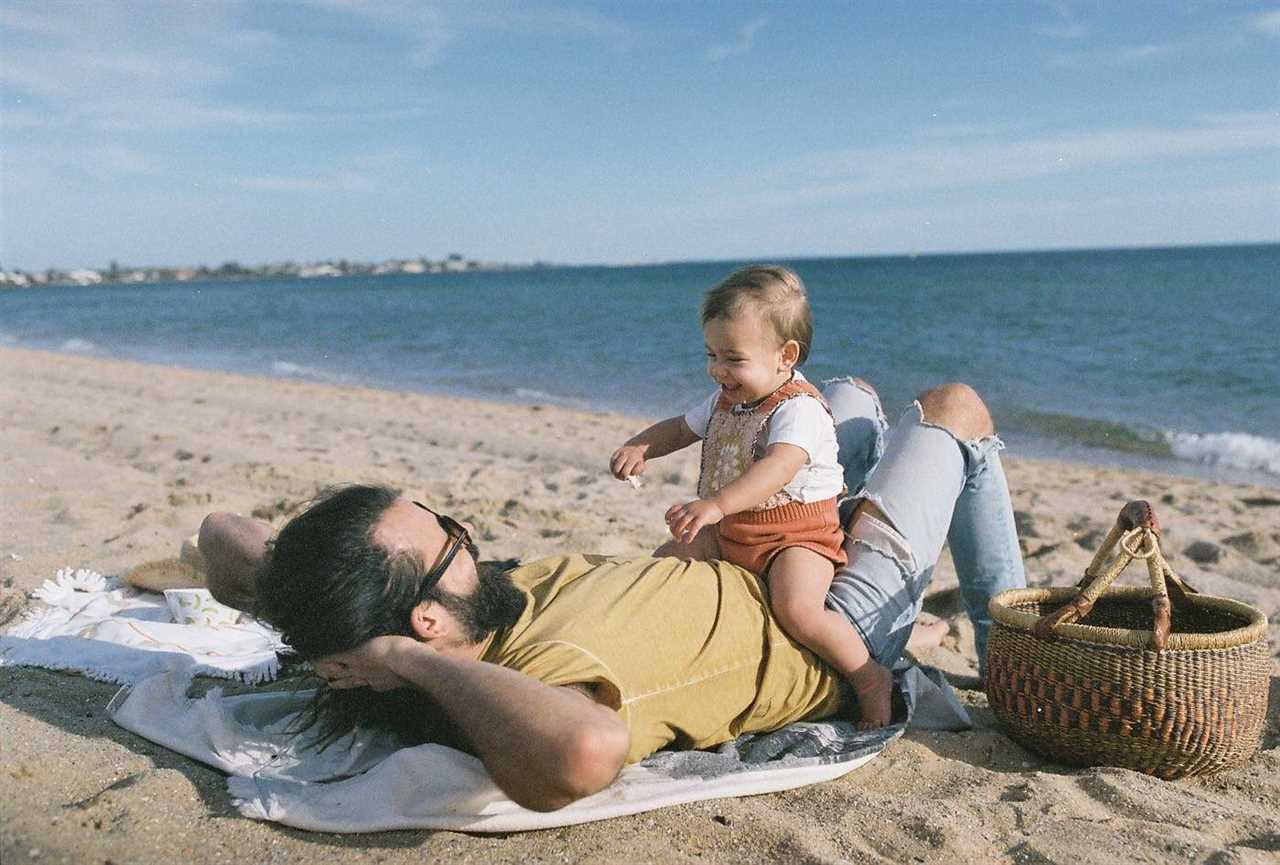 Husband and baby daughter sit on top of him while they are on the beach for a story about where new dads can find support.