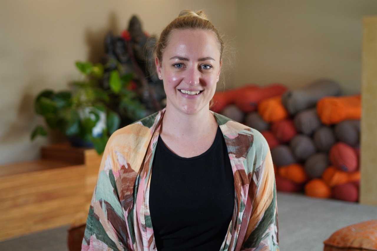 A woman with her hair pulled back into a bun wearing a black top and multi-coloured jacket stands in front of rolled yoga mats