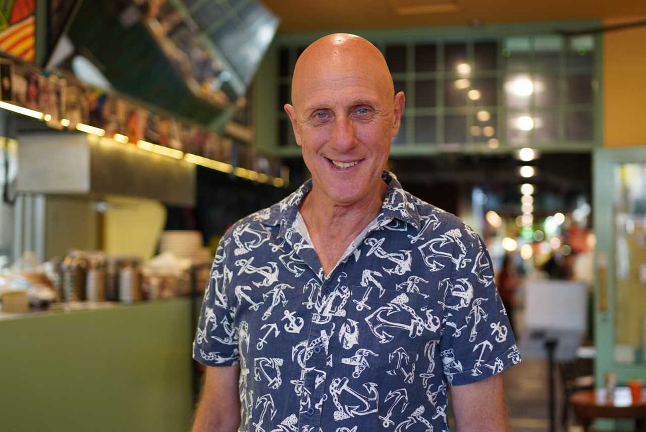 A man in a navy and white patterned button up shirt stands inside a cafe, smiling