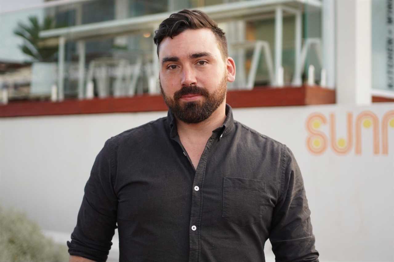 A man wearing a black button up shirt with brown hair and a beard stands in front of a restaurant